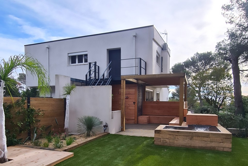 Aménagement complet de jardin | Espace Zen sur mesure. Bassin traverse bois, terrasse et pergolas en bois. Gazon synthétique. Composition de végétaux adaptés au climat sec, nécessitant peu d’arrosage. Implantation d’un olivier. 